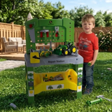 A young child stands next to a green John Deere-themed children's repair station playset on a grassy backyard lawn. The green workbench features a simulated engine bay, a yellow storage bin, and toy tractors and tools.