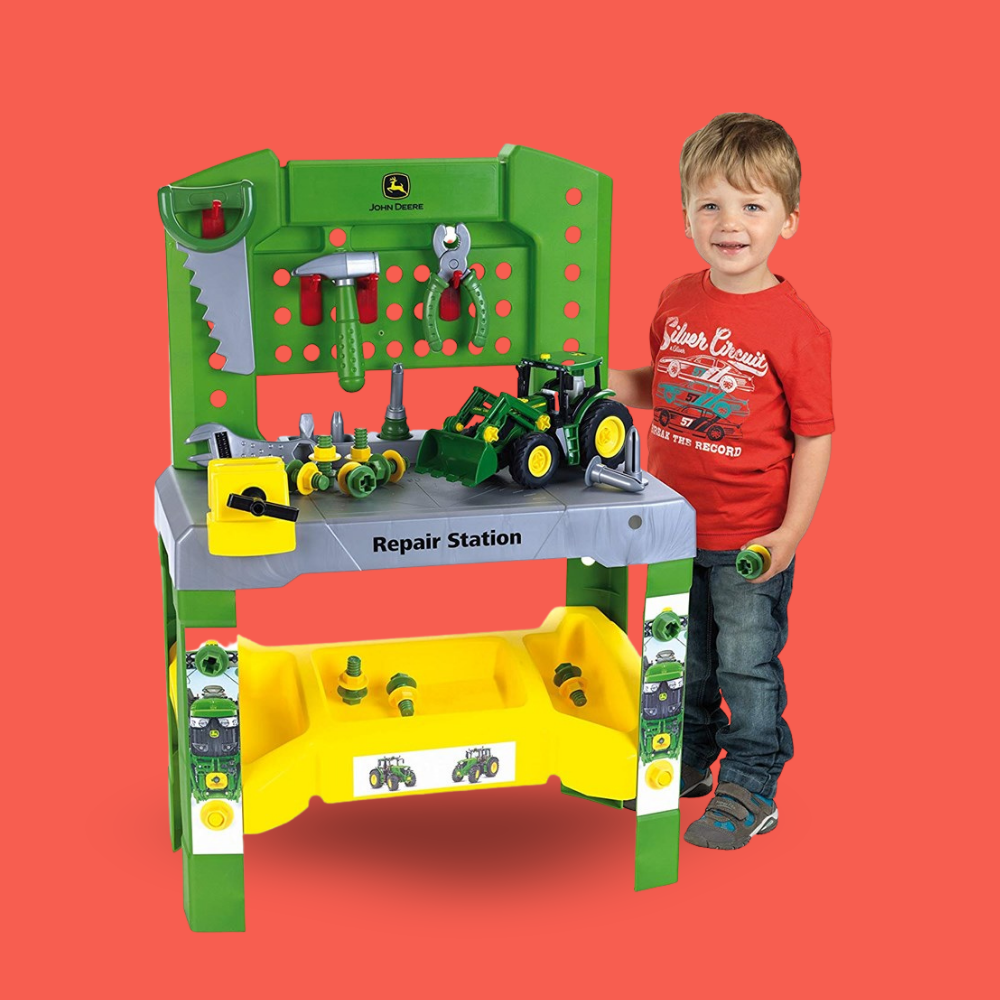A young boy stands next to a green John Deere wooden toy repair station playset, holding a small green toy tractor, against a solid red background.