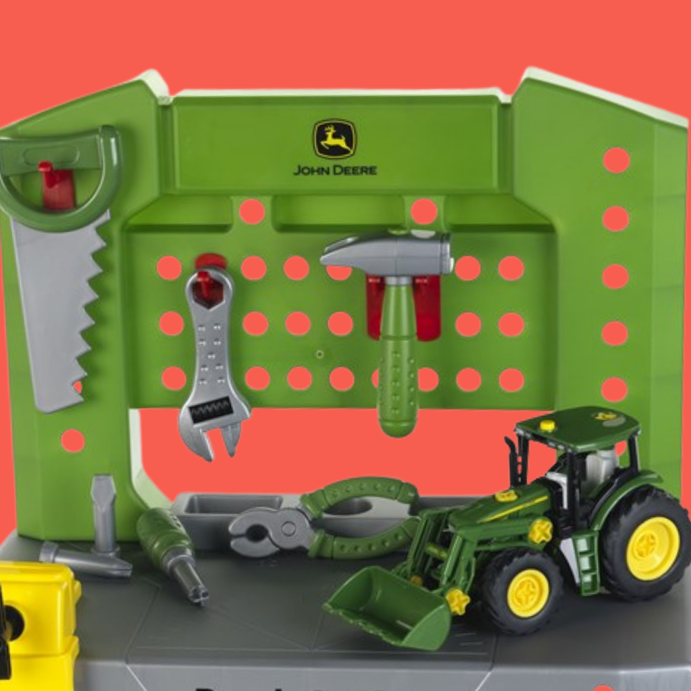 John Deere toy workbench playset with green pegboard, hanging tools, and a green tractor with yellow wheels on a gray base against a coral background.