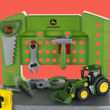 John Deere toy workbench playset with green pegboard, hanging tools, and a green tractor with yellow wheels on a gray base against a coral background.
