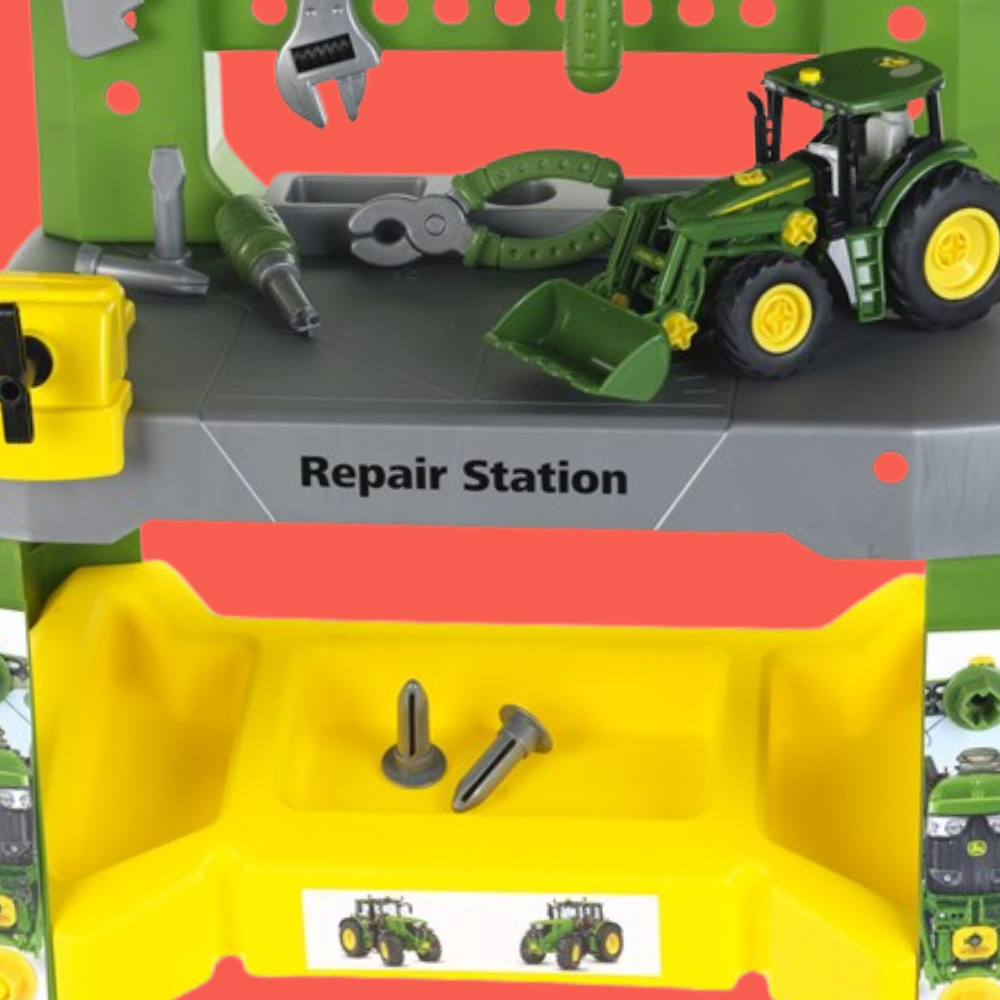 A green John Deere toy tractor with a front loader sits on a gray workbench inside a colorful toy repair station playset. The playset features a green upper section with hanging tools, a gray workbench labeled 'Repair Station,' and a bright yellow lower storage compartment with screws.