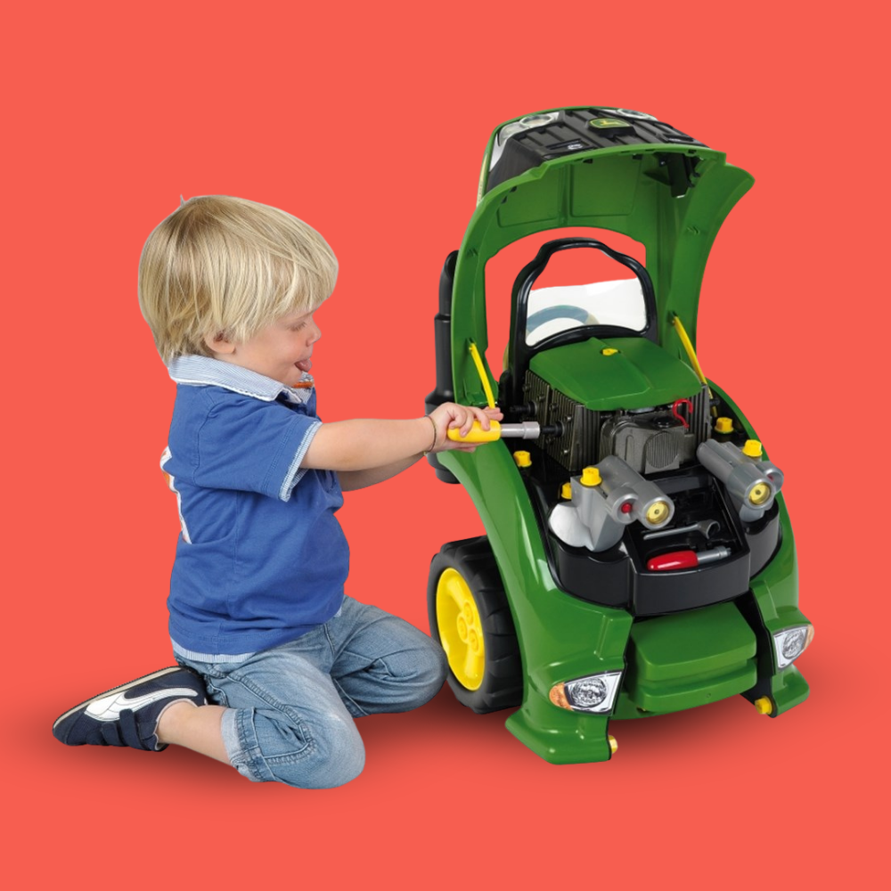 A young blonde boy in a blue shirt and jeans kneels beside a green toy tractor with an open hood, reaching toward the engine area against a solid red background.