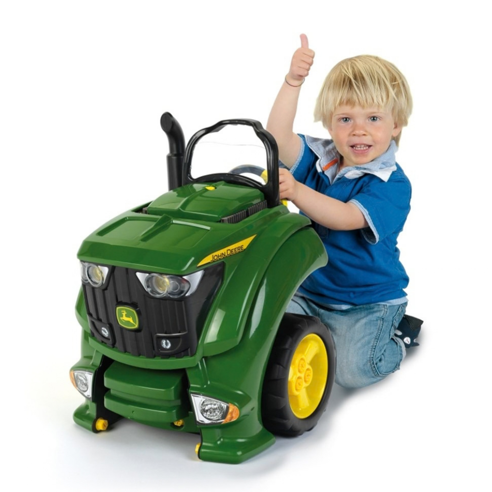 A young blonde boy in a blue polo shirt and jeans rides a green John Deere toy tractor with yellow wheels, giving a thumbs-up against a white background.