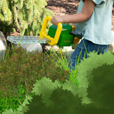 A child wearing a light blue shirt and jeans is using a green and yellow toy chainsaw to cut green grass and bushes in an outdoor garden setting.