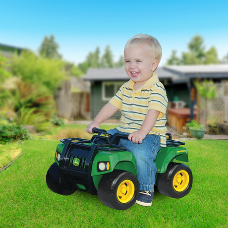 A smiling toddler wearing a yellow striped polo shirt and jeans sits on the John Deere Buck ATV ride-on toy, riding on a lush green lawn with a backyard and a small house in the background.