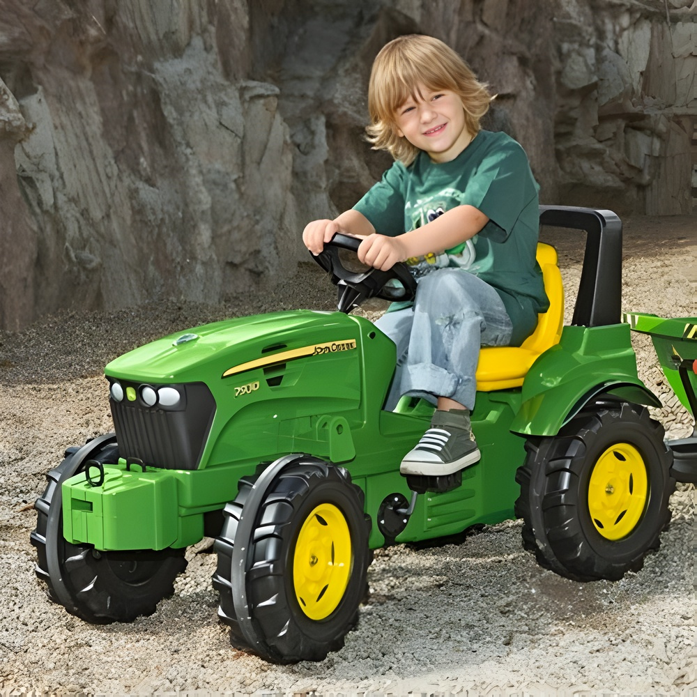 Child sitting on a green toy tractor with yellow wheels in front of a stone wall.