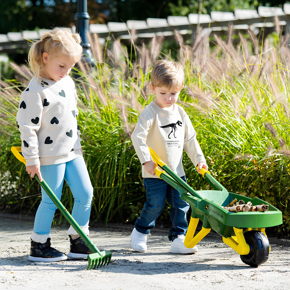 Two children outdoors in a garden setting, one holding a green rake and the other pushing a green and yellow wheelbarrow filled with small stones.