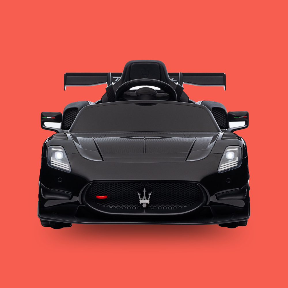 A black Maserati-style children's ride-on toy car shown from the front on a solid coral-red background. The car features a glossy black finish, a front grille with the Maserati trident emblem, headlights, side mirrors, and a rear wing.