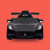 A black Maserati-style children's ride-on toy car shown from the front on a solid coral-red background. The car features a glossy black finish, a front grille with the Maserati trident emblem, headlights, side mirrors, and a rear wing.