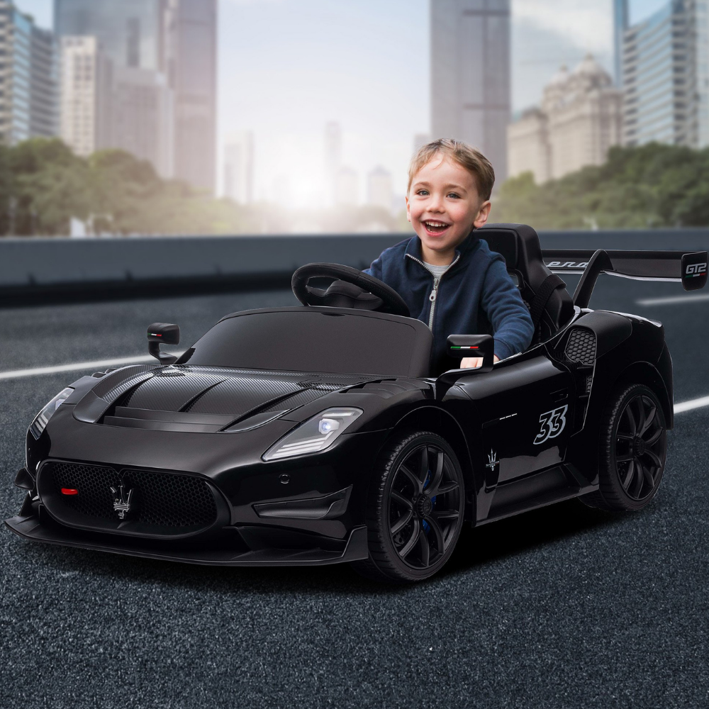 A young boy smiling and driving a black Maserati-themed toy sports car on a road with a blurred city skyline background.