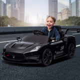 A young boy smiling and driving a black Maserati-themed toy sports car on a road with a blurred city skyline background.