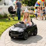 A young girl wearing a party hat drives a black toy car designed like a modern SUV on a paved backyard path. A man grills on a charcoal grill in the background, and other children and balloons are visible in the yard.