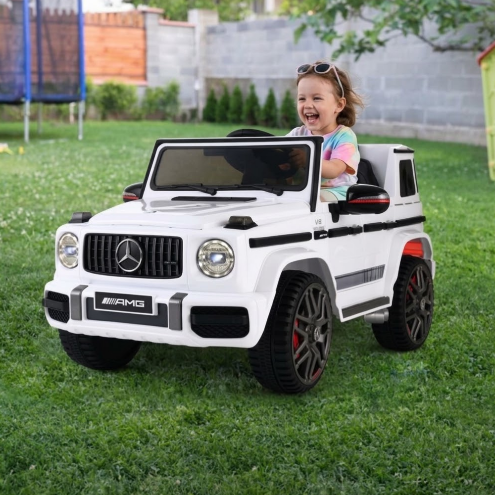 A young child wearing sunglasses sits smiling in a white Mercedes-Benz AMG ride-on toy car on a green grassy backyard lawn.