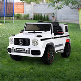 A white Mercedes-Benz G-Class style children's ride-on toy car parked on a green grass lawn, with a backyard fence and trampoline visible in the background.