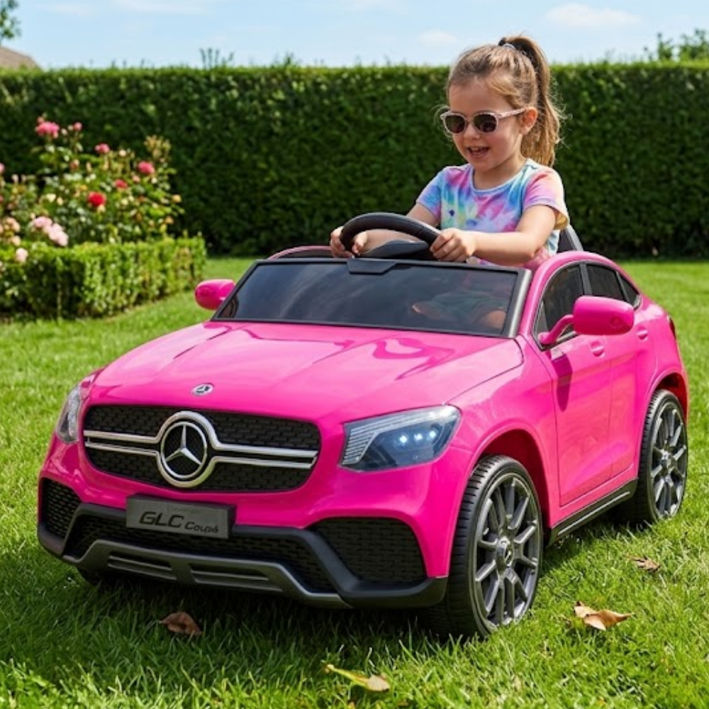 A young girl wearing sunglasses and a tie-dye shirt drives a bright pink Mercedes-Benz GLC toy car on a green grassy lawn with a hedge in the background.