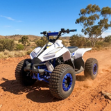 A white and blue Yamaha quad bike ATV with large off-road tires, shown driving on a dusty red dirt track in an outdoor desert landscape under a clear blue sky.