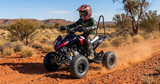 A person wearing a red and black helmet rides a red and black quad bike ATV on a dusty red dirt road through an arid desert landscape.
