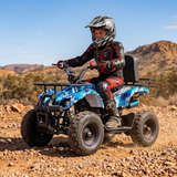A child wearing a black, red, and gray motocross helmet and suit rides a blue and black quad bike on a rocky desert trail with mountains in the background.