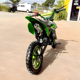 Green dirt bike parked on a concrete surface with vehicles and trees in the background