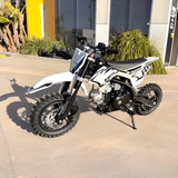 White and black dirt bike parked on a concrete surface with plants and a building in the background.