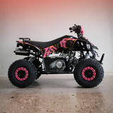 A small black ATV with bright pink and yellow graphic decals, featuring large knobby tires with pink rims, photographed against a plain indoor wall with a warm gradient.