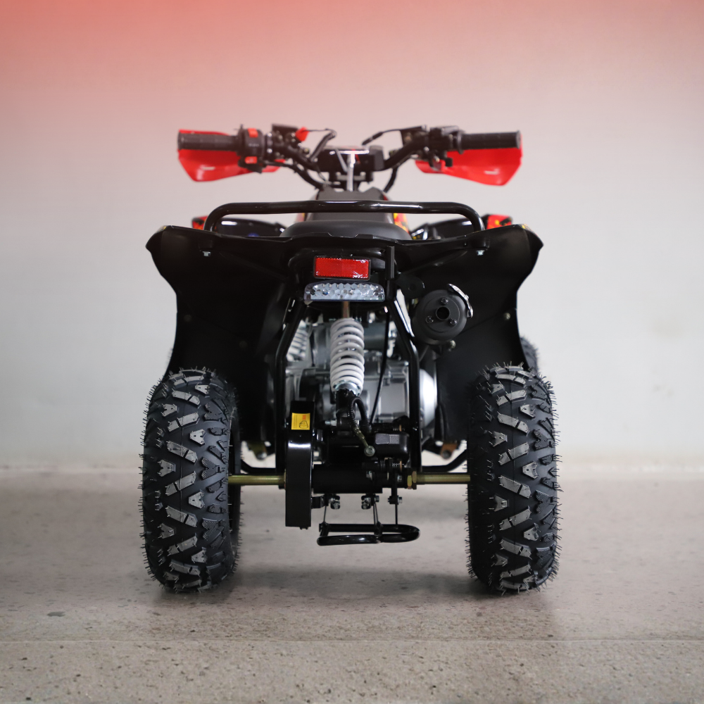 Rear view of a black quad bike ATV with red handlebar grips and a silver engine, parked on a concrete floor against a light gray wall.