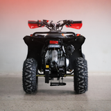 Rear view of a black quad bike ATV with red handlebar grips and a silver engine, parked on a concrete floor against a light gray wall.