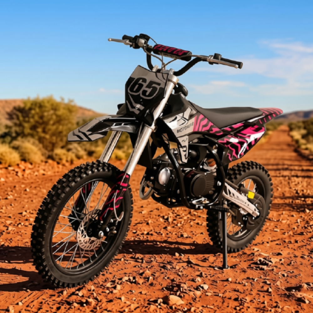 A pink and black dirt bike with the number 65 on its front plate, parked on a red dirt road with a blurred outdoor desert background.