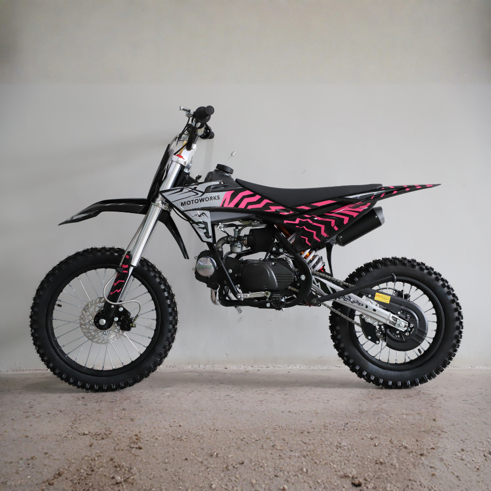 A mini dirt bike with a black and pink zebra-stripe pattern, photographed from the side against a plain light gray wall and concrete floor.