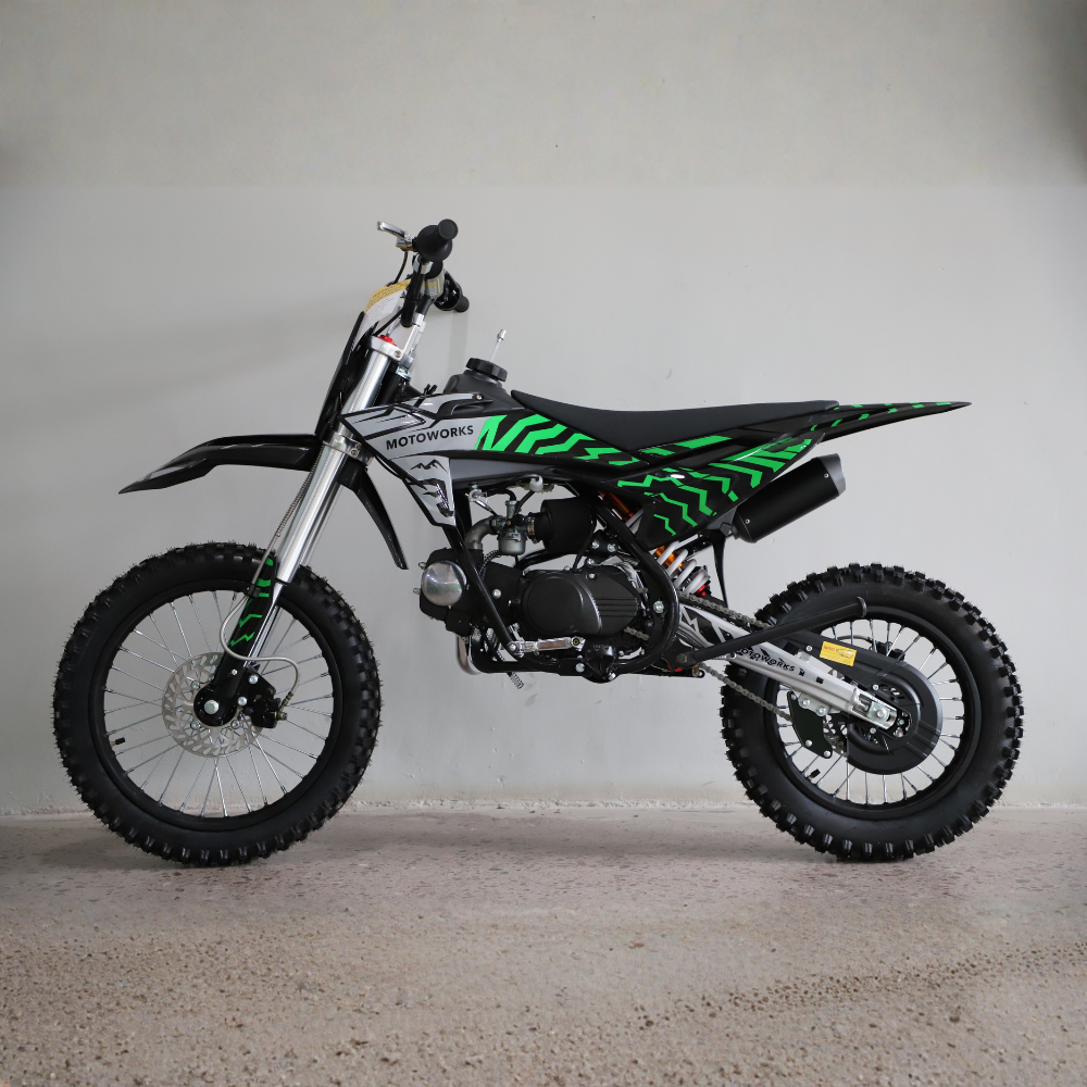 A small off-road dirt bike with a black seat and bodywork, featuring bright green graphic stripes, photographed in profile against a plain light gray wall and concrete floor.