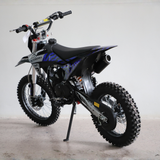 A black and blue off-road dirt bike with knobby tires, a black seat, and a visible exhaust pipe, photographed indoors against a plain light gray wall.