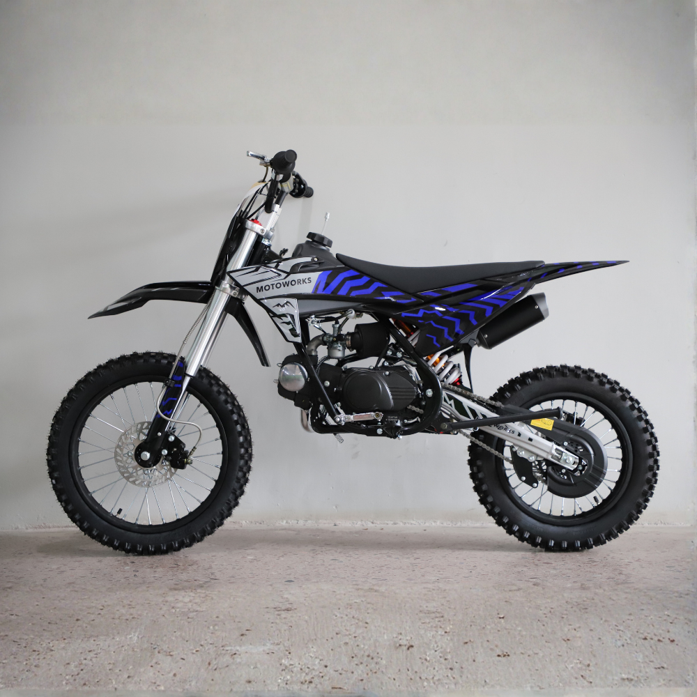 A small off-road dirt bike with a black frame, black seat, and blue graphic decals, photographed in profile against a plain light gray wall and concrete floor.