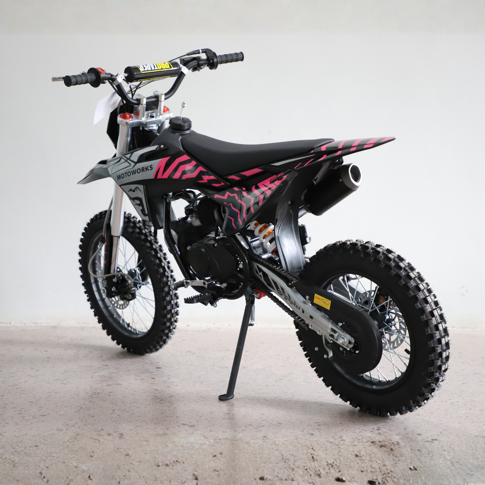 A black and pink mini dirt bike with knobby tires, parked on a concrete floor against a plain light gray wall.