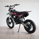 A black and pink mini dirt bike with knobby tires, parked on a concrete floor against a plain light gray wall.