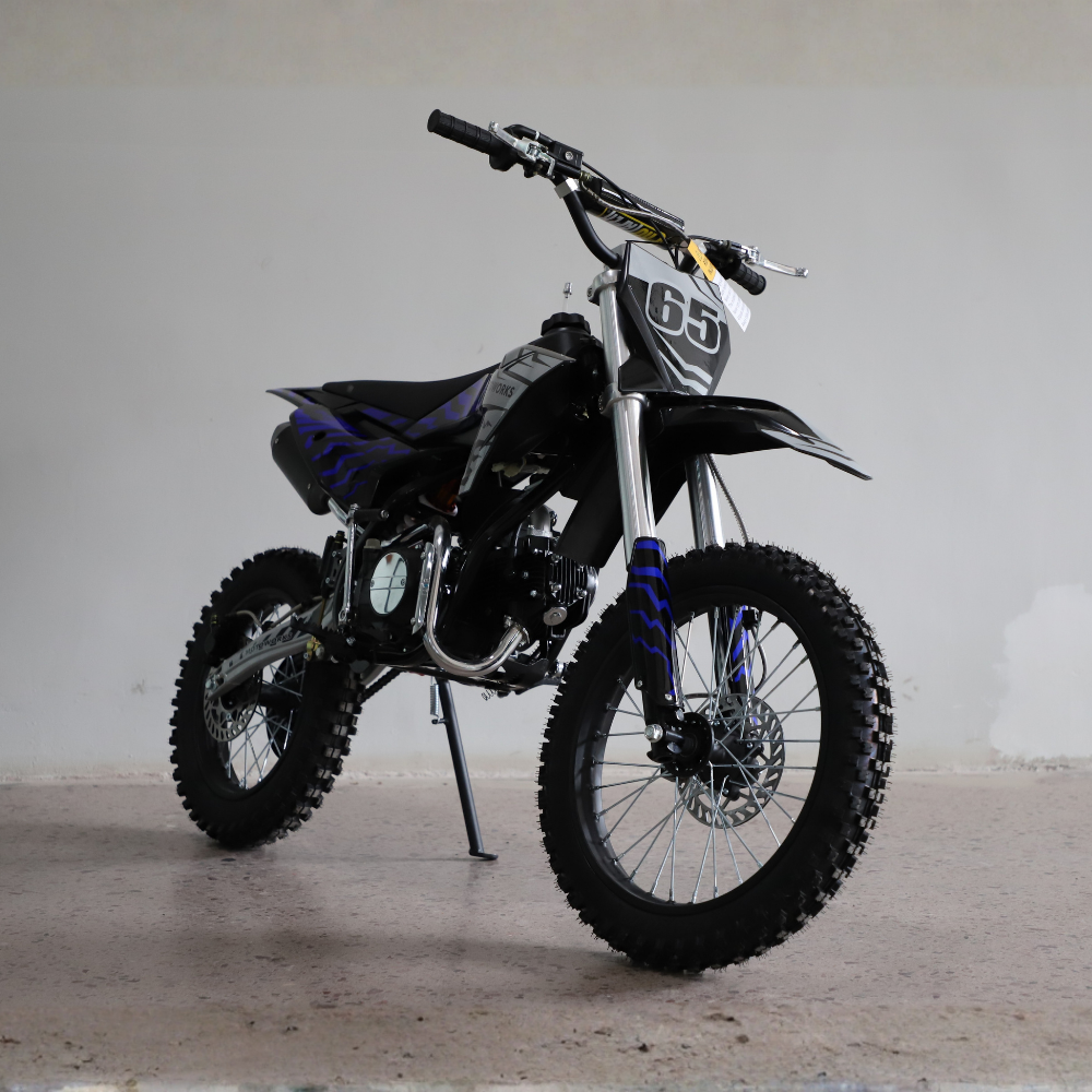 A black and silver dirt bike with blue flame decals and a white number 65 plate, photographed on a concrete floor against a plain light gray wall.