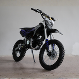 A black and silver dirt bike with blue flame decals and a white number 65 plate, photographed on a concrete floor against a plain light gray wall.