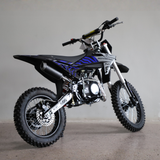 A black and blue dirt bike with a silver engine and black knobby tires, photographed from a three-quarters angle against a plain gray studio background.