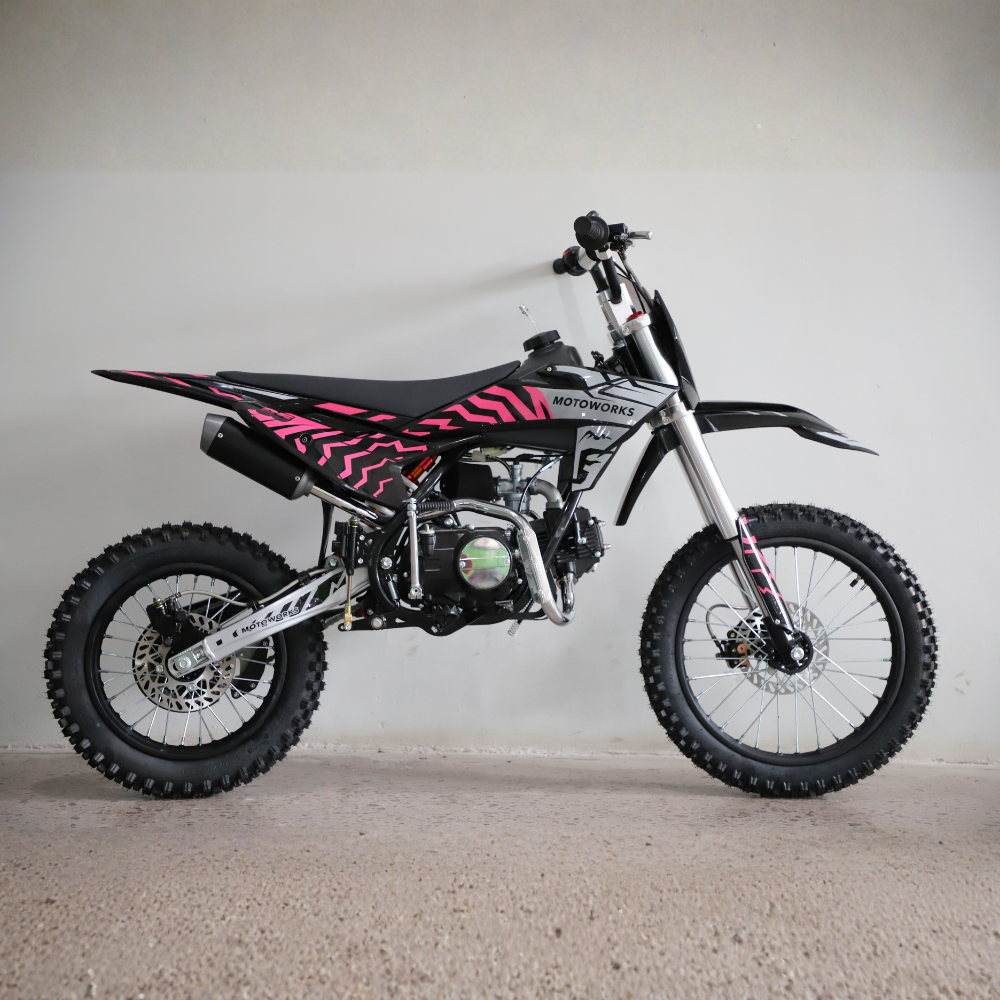 A small off-road dirt bike with a black and pink geometric pattern on the side panels, photographed in profile against a plain light gray wall and concrete floor.
