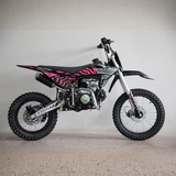 A small off-road dirt bike with a black and pink geometric pattern on the side panels, photographed in profile against a plain light gray wall and concrete floor.