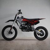 A small off-road motorcycle with a black and red patterned seat, black fenders, and a silver engine, photographed in profile against a plain light gray wall.