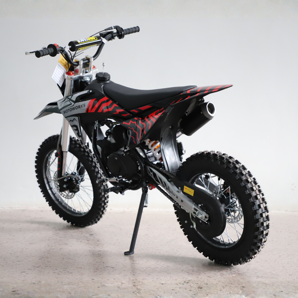 A small off-road dirt bike with knobby tires, a black seat, and red and black graphic decals, photographed on a light gray studio floor with a white backdrop.