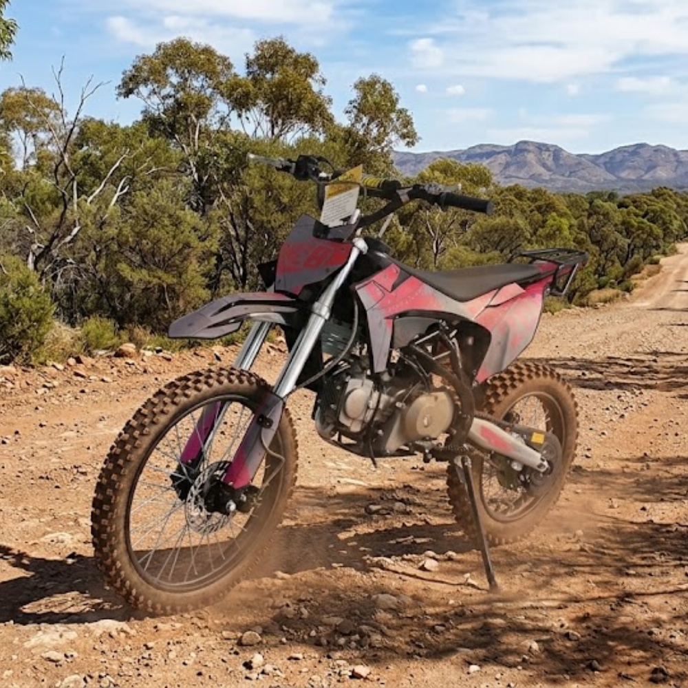 A red and black dirt bike motorcycle is shown parked on a dusty dirt road in an outdoor desert landscape with trees and mountains in the background.