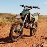 A green and black dirt bike with knobby off-road tires parked on a reddish-brown dirt trail under a clear blue sky.