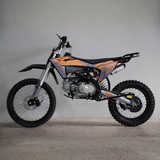 A dirt bike with orange and gray graphics, black seat, and silver engine components, photographed in profile against a plain gray wall and concrete floor.