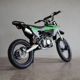A green and black mini dirt bike with a silver exhaust and black knobby tires, photographed on a concrete floor against a plain gray wall.