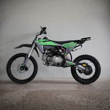 A small off-road dirt bike with green and gray graphics, black seat, and knobby tires, photographed in profile against a plain concrete wall and floor.