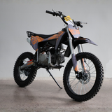 A small off-road dirt bike with a black and orange color scheme, standing on its kickstand in a studio setting with a light gray background.