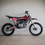 A red and black mini dirt bike with knobby tires, parked against a plain light gray wall on a concrete floor.