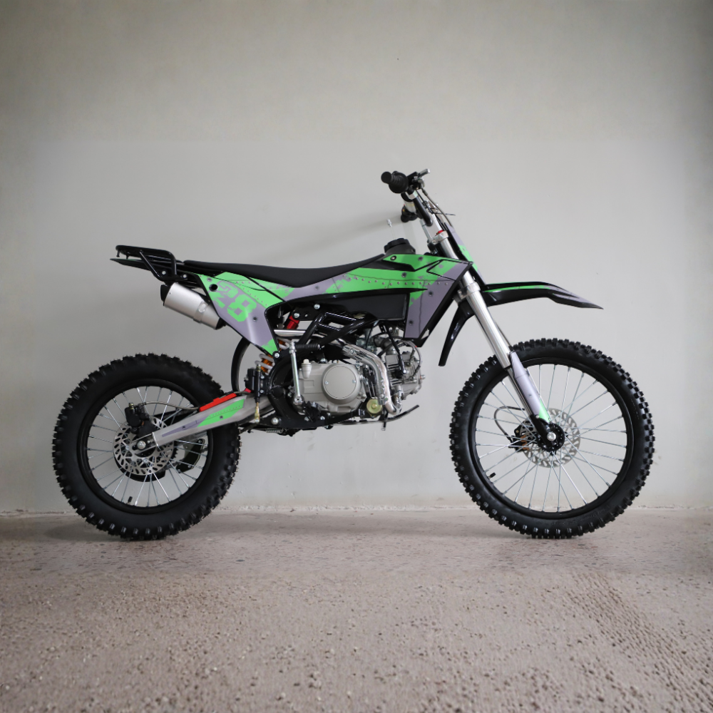 A green and black mini dirt bike with a silver exhaust and knobby tires, photographed in profile against a plain light gray wall and concrete floor.