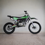 A green and black mini dirt bike with a silver exhaust and knobby tires, photographed in profile against a plain light gray wall and concrete floor.